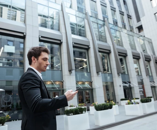 man looking at phone with building behind -commercial real estate loans